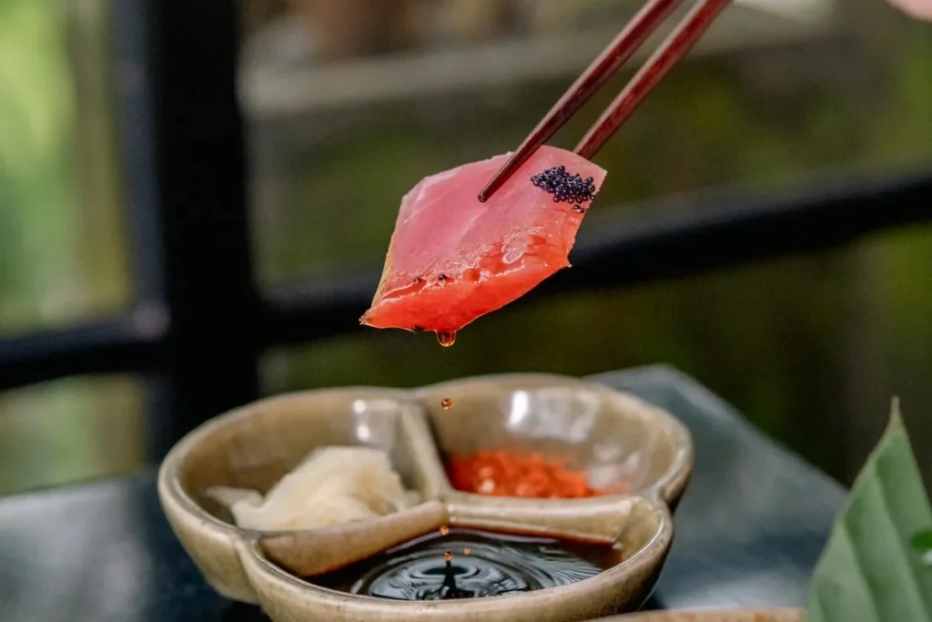 Piece of sashimi held with chopsticks above dipping sauce for Japanese dining.