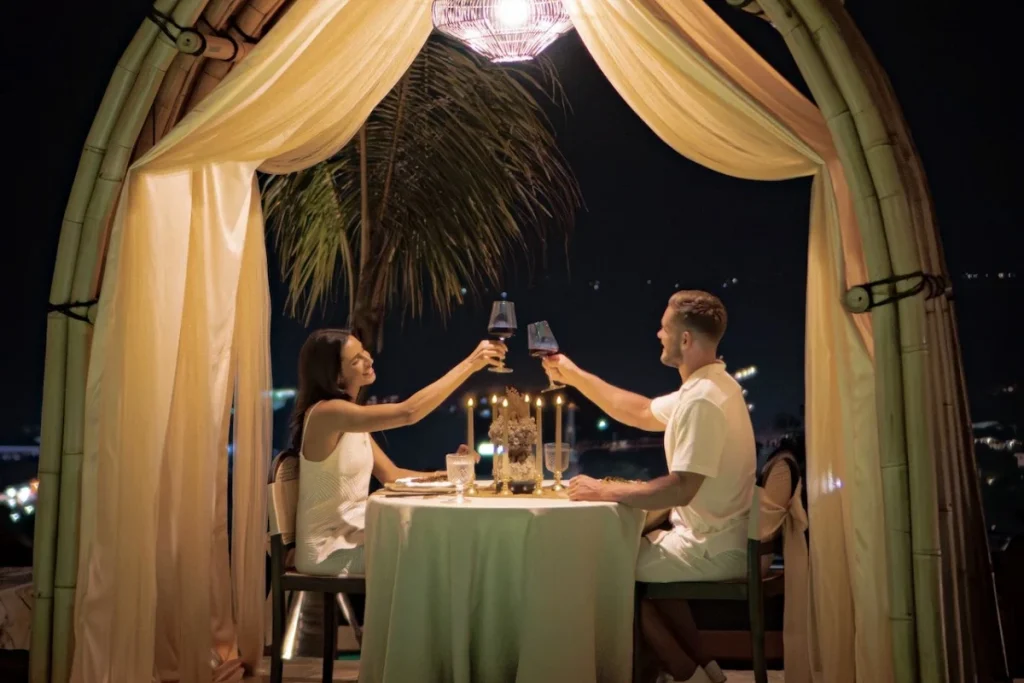 A couple toasts with wine during a romantic anniversary dinner under a bamboo canopy at night in Bali.