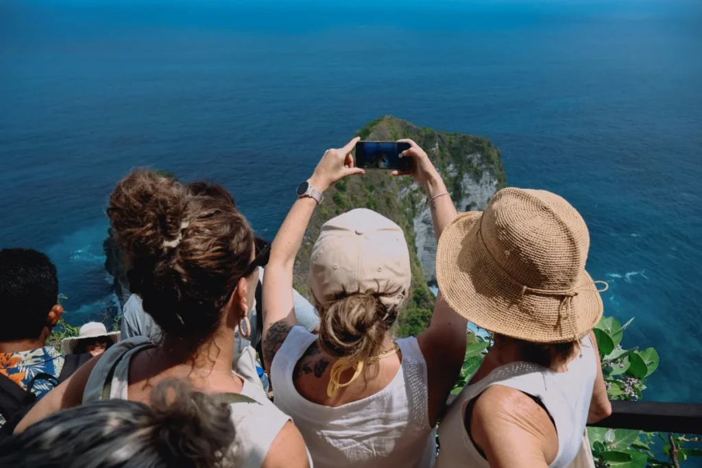 Travelers photograph a dramatic Bali cliff surrounded by deep blue ocean from a busy coastal viewpoint.