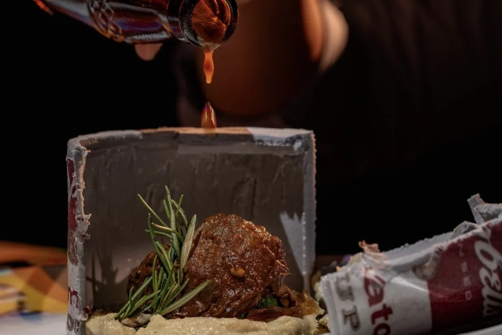 A plated meat dish with rosemary is served as part of a tasting menu, while sauce is poured over it.