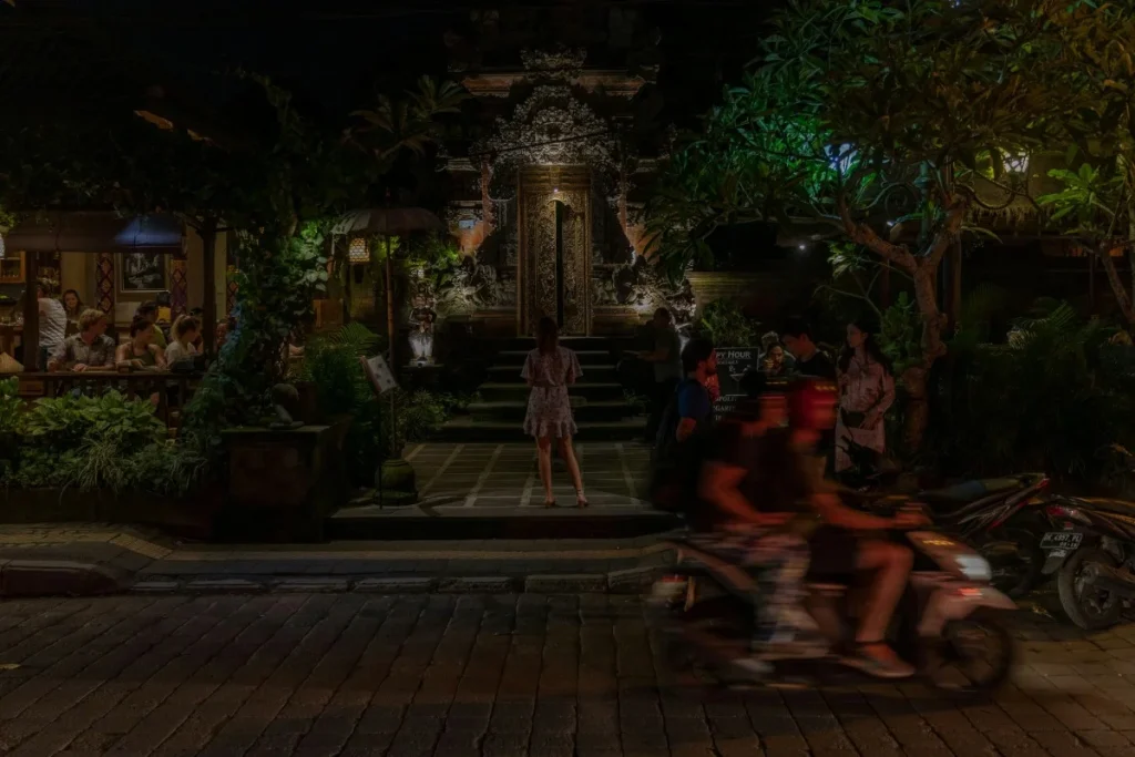 A busy Ubud street at night shows a temple entrance, diners, pedestrians, and motorbikes passing by.