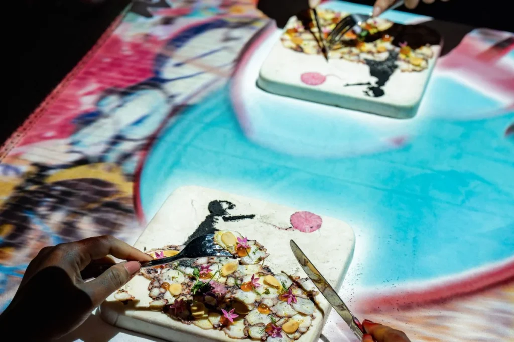A guest uses a fork to enjoy plated food while colorful projections create an immersive dining experience in Bali.